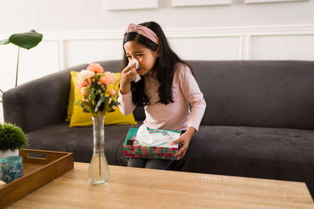 Sick cute girl blowing her nose and holding a box of tissues while sitting in the living room