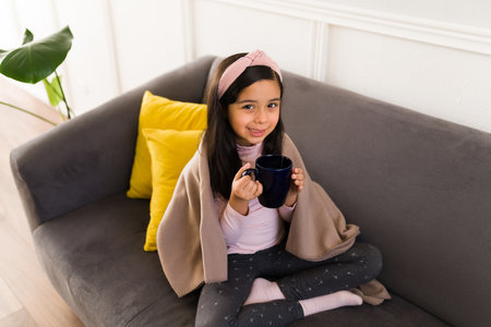 Happy little girl relaxing on a leisure day and drinking a hot tea while wrapped in a blanketの写真素材