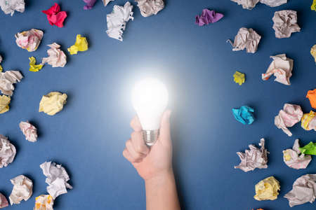 Young adult having a new bright idea surrounded with discarded papers while holding a lit lightbulb against a blue backgroundの写真素材