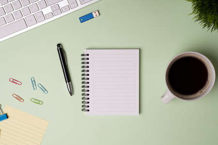 Office workspace. Desk with a computer keyboard, open notepad, pen and paper clips next to a cup of...