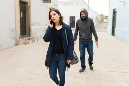 Insecure city. Beautiful woman walking alone in a street while talking on the phone. Young woman being chased by a dangerous thiefの写真素材