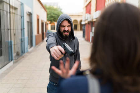 Violent hispanic man and thief pointing a gun to a young woman to rob her money and her smartphone on an alleyの写真素材