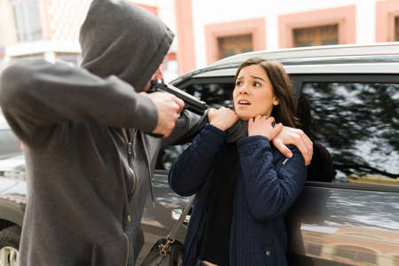 Aggressive young man and dangerous criminal pointing a gun to a scared caucasian woman to steal her carの写真素材