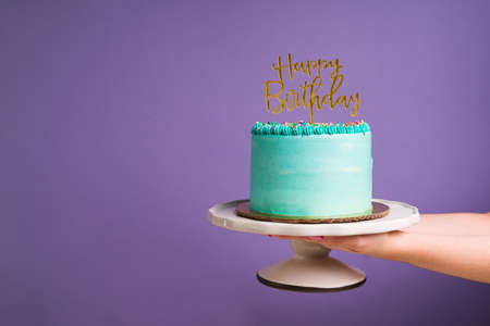 Woman's hand holding a cake with a gold happy birthday sign in front of a purple background copy...