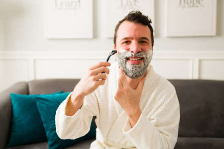 Excited man in a white robe smiling while using shaving cream after getting out of the showerの写真素材