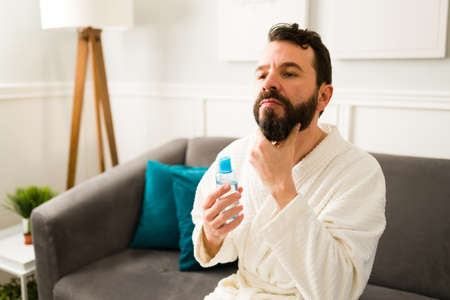 Good-looking man using a beard treatment and oil for a beautiful shiny hair while relaxing at homeの写真素材