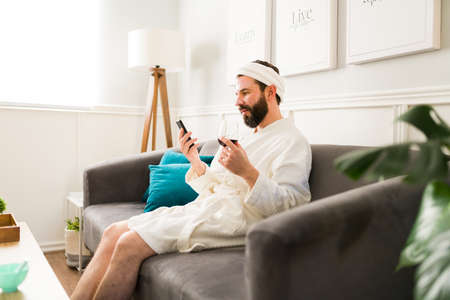 Beauty spa day. Young man with a face mask texting on his smartphone while having a relaxing day with a glass of wineの写真素材