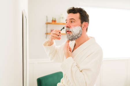 Handsome man getting out of the shower on a white robe and looking in the mirror while shaving his...