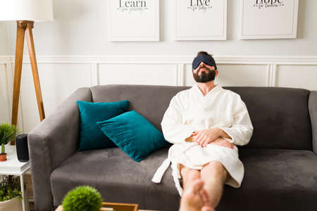 I'm so tired. Relaxed latin man with a white robe resting on the sofa with a sleeping mask