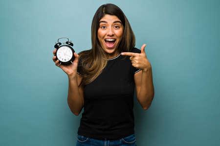Daylight saving time. Beautiful hispanic woman showing an alarm clock and looking happy about the time changeの写真素材