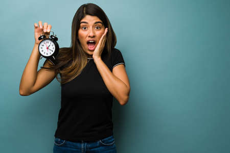 It's very late. Shocked young woman showing an alarm clock and looking surprised by the late hour