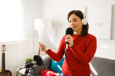 Beautiful young woman singing a song with a microphone while reading the lyrics. Singer recording new music at homeの写真素材
