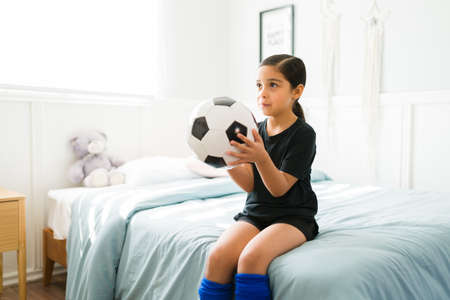 Adorable little child holding a ball while sitting on her bed and ready to go to a football soccer practiceの写真素材