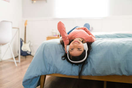 I love this song. Happy young girl smiling and lying upside down in bed while listening to music with headphonesの写真素材
