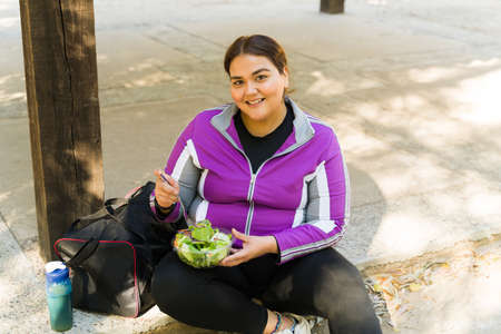 Portrait of a beautiful plus size woman looking at the camera and smiling while eating a green salad...