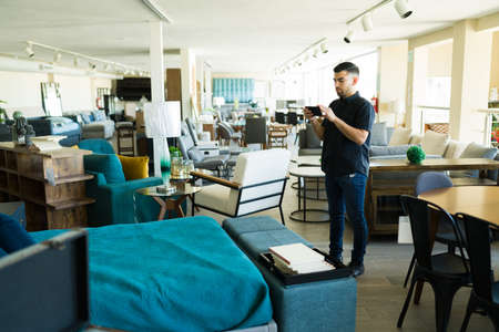 Handsome man taking pictures of the beautiful furniture and texting at the store while shopping for...
