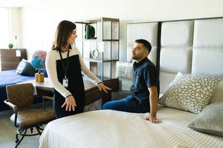 Beautiful sales representative speaking with a young man and trying a comfortable bed at the furniture storeの写真素材