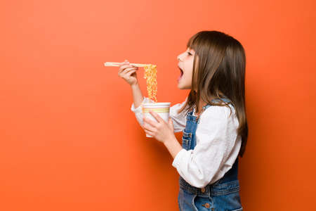 Profile view of a little girl opening a big mouth and eating noodles with chopsticks in a studioの写真素材