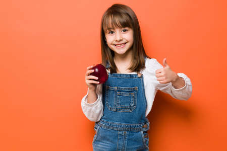 Cute little girl holding an apple in one hand and making a thumb up in a studioの写真素材