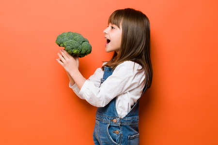 Profile view of a little brunette girl eating some broccoli in a studio with her mouth wide openの写真素材