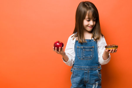 Cute little girl with her tongue out wanting to eat a donut with sprinkles instead of going fot the healthy choice of an appleの写真素材
