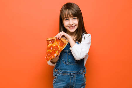 Portrait of a beautiful Caucasian girl holding a big slice of pizza and feeling really hungry in a...