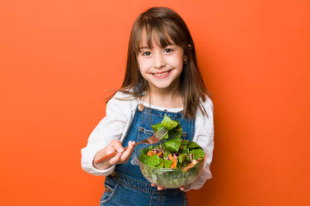 Good looking little girl eating a salad from a bowl and making eye contact in a studioの写真素材