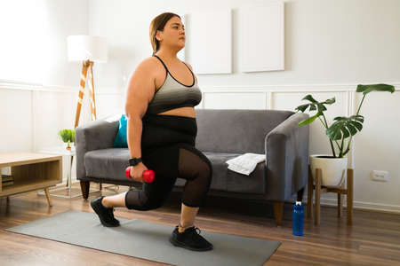 Motivated fat woman doing lunges while using dumbbell weights to try to lose some weight for a healthy lifestyleの写真素材