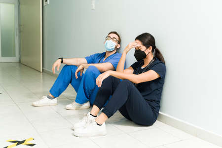 Exhausted nurse and doctor with face masks resting in the hospital aisle. Medical staff feeling tiredの写真素材