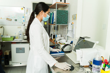 Concentrated female biochemist with a face mask making medical research at the laboratory with blood...