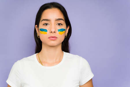 Closeup of a young woman with a Ukranian flag painted on her face while looking serious in a studio