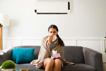 Sick young woman blowing her nose and feeling ill because of the cold air flow of the air conditionerの写真素材