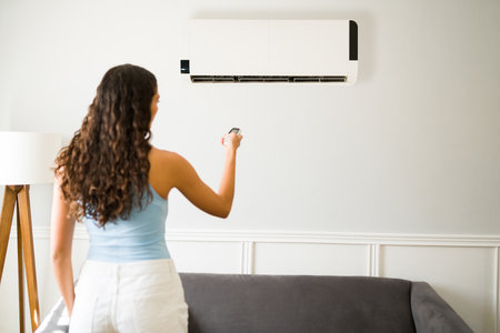 Hispanic woman seen from behind turning on her air conditioning with a remote during a heat waveの写真素材