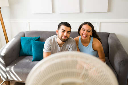 Attractive smiling couple enjoying the air from the electric fan and cooling down because of the...