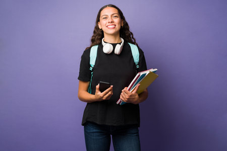 College student holding her smartphone and books while carrying a backpack against a studio purple backgroundの写真素材
