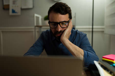 Depressed man wearing glasses and typing on the laptop while working from home very late