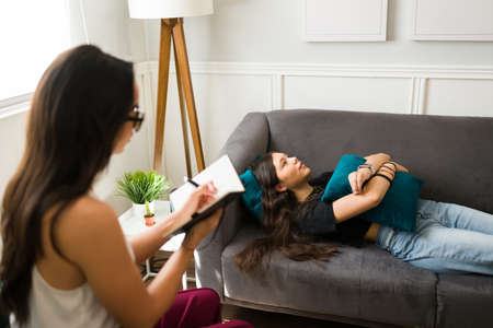 Rear view of a young female psychologist taking notes and giving mental health advice to a teen patientの写真素材