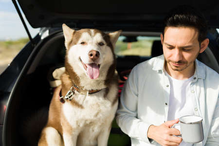 Thoughtful young man drinking coffee and resting while taking a vacation trip by car with his husky...