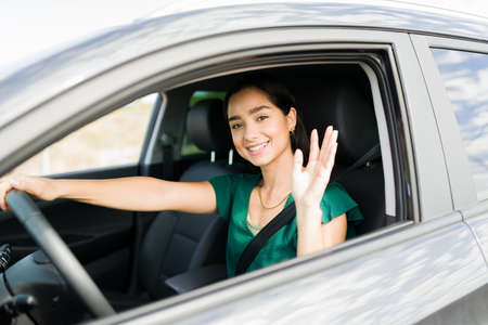 Happy young woman waving hello and smiling while driving her new beautiful car