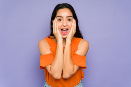 Attractive young woman with her hands on the face smiling and looking surprised while screamingの写真素材
