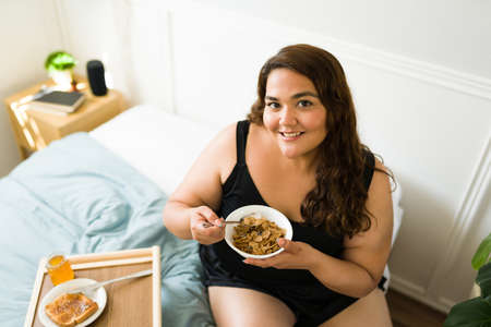 High angle of woman with obesity looking at the camera while enjoying breakfast in bed and coffee...
