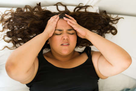 Stressed obese woman rubbing her head while suffering from a bad migraine or headache in bedの写真素材
