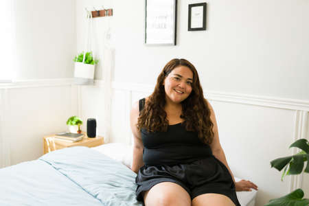 Cheerful big young woman sitting in bed. Hispanic obese woman enjoying a leisure day in her beautiful bedroomの写真素材