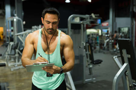 Strong muscular coach writing a workout routine and fitness plan on a clipboard at the gym