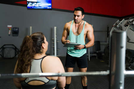 Athletic personal trainer giving a workout routine and diet plan to an overweight young woman at the gymの写真素材