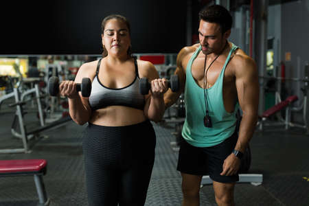 Determined overweight woman in activewear lifting dumbbell weights and doing a strong workout with a personal trainerの写真素材