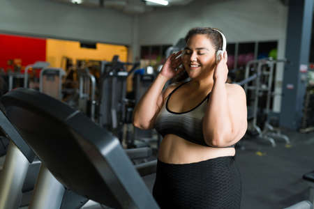 Excited plus size woman listening to music with wireless headphones while running on the treadmill at the gymの写真素材