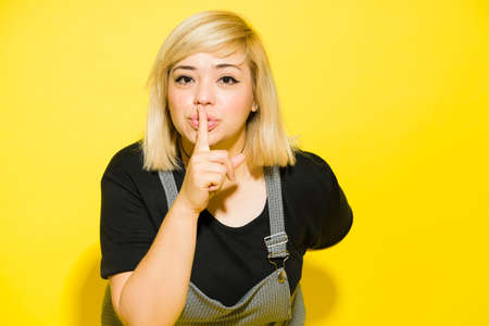 Big Caucasian woman trying to keep a secret with a finger on her lips in a studio with yellow...