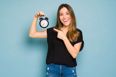 Attractive woman in casual clothing pointing at an alarm clock and smiling in a studioの写真素材