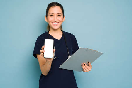 Young female doctor showing a mobile app on her smartphone screen and smiling in a studioの写真素材